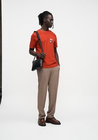 Κόκκινο βαμβακερό μπλουζάκι με μικρό λογότυπο, μπεζ παντελόνι ραμμένο στα μέτρα, και καφέ δερμάτινα παπούτσια, συμπληρωμένα με μια μαύρη τσάντα χιαστί.