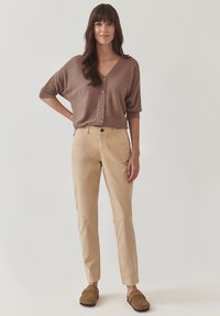 Hnědý svetr s V výstřihem a knoflíky, kombinovaný se světle béžovými kalhotami rovného střihu. Model nosí hnědé boty na slip-on. Jednoduchý a uvolněný střih.