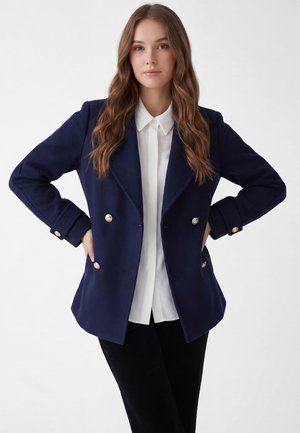 Woman with wavy brown hair wearing a navy double-breasted blazer with gold buttons over a white collared shirt, posing with hands on hips.