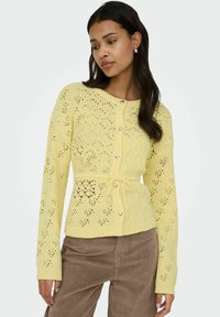 Cardigan tricoté jaune clair avec des motifs en dentelle complexes, des boutons en forme de cœur et une ceinture nouée à la taille, associé à un pantalon en velours côtelé marron.