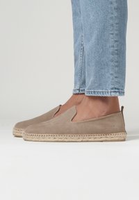 Espadrilles en suède brun clair avec ouvertures latérales élastiques, semelle en jute et marquage minimal au talon. Jeans visibles au-dessus de la chaussure.