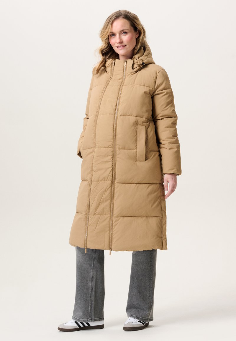 Vrouw in een lange beige gewatteerde jas met capuchon, grijze spijkerbroek en witte sneakers met zwarte strepen, staande tegen een effen achtergrond.