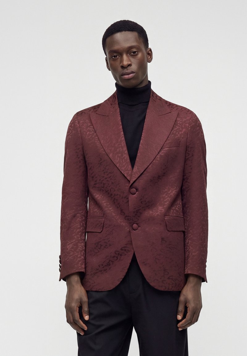 Homme portant un blazer rouge foncé texturé par-dessus un col roulé noir et un pantalon noir, debout devant un fond gris clair uni.