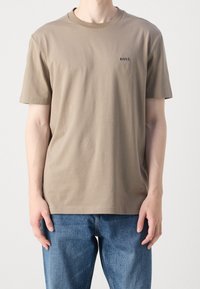 Hnědé tričko s krátkým rukávem vyrobené z bavlny, s kulatým výstřihem a malým černým logem na levé hrudi. Nosené s modrými džíny.