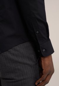 Chemise noire à manches longues avec des poignets boutonnés, tissu lisse, coupe ajustée et détails d'épaules affûtés, associée à un pantalon en gris foncé texturé.