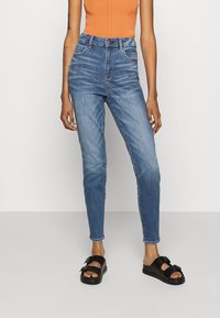 Högmidjade blå denimjeans med smal passform, med två framfickor och subtila blekningar. Kombineras med svarta sandaler och en orange topp.