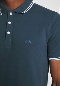 Tmavě zelená polo košile s texturovanou látkou, klasickým límečkem s bílými pruhy a třemi knoflíky. Vyšitá logo na hrudi.