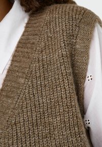 Bruine gebreide vest met een geribbelde textuur en brede armsgaten. Gelaagd over een wit shirt met perforaties op de mouwen.