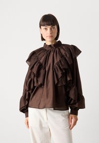 Brun flæset bluse med høj krave, strukturerede ærmer og blød tekstur. Modellen bærer hvide ribbede bukser. Enkel, elegant design.