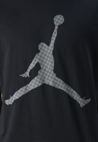 Sort t-shirt med en stor grå silhuet af en hoppende figur, der holder en basketball, med et struktureret mønster gennem hele designet.