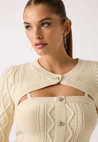 Top lavorato a maglia color crema con motivi a treccia, caratterizzato da un collo rotondo, un design con aperture e dettagli in argento sui bottoni.