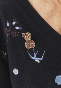 Brązowa broszka w kształcie misia z emalii z złotym obramowaniem, przypięta do granatowego wełnianego ubrania z kolorową, kwiatową haftowaną dekoracją i wzorami.