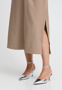 Vestido midi beige con abertura lateral, combinado con tacones plateados brillantes de punta afilada que cuentan con una delgada correa en el tobillo y un tacón pequeño.