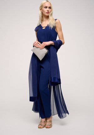 Tuta blu navy con sovrapposizione trasparente blu navy, caratterizzata da un collo a cuore e sandali con dettagli dorati. Clutch grigia con finitura metallica.