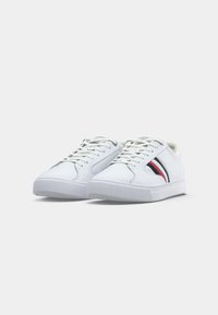 Pantofi sport albi din material sintetic, cu o talpă din cauciuc texturat. Dungi roșii și albastre notabile pe lateral și un design clasic cu șireturi.