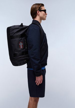 Black nylon backpack with PAREPURI logo and Norwegian flag detail. Model wearing navy bomber jacket and shorts, with sunglasses, viewed from the side.