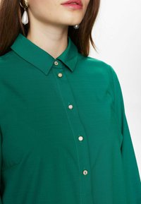 Camicia verde con bottoni, caratterizzata da un colletto classico, una texture liscia e bottoni decorativi in tono oro lungo il pattino anteriore.