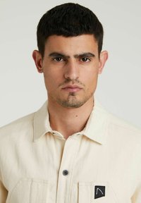 Camicia beige con bottoni, realizzata in cotone, dotata di due tasche frontali, colletto e una toppa nera con logo sulla tasca sinistra. Texture liscia.