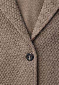 Taupe gebreide blazer met reliëfstructuur, voorzien van ingesneden revers en een enkele donkere knoop met gedeeltelijk zichtbare letters.