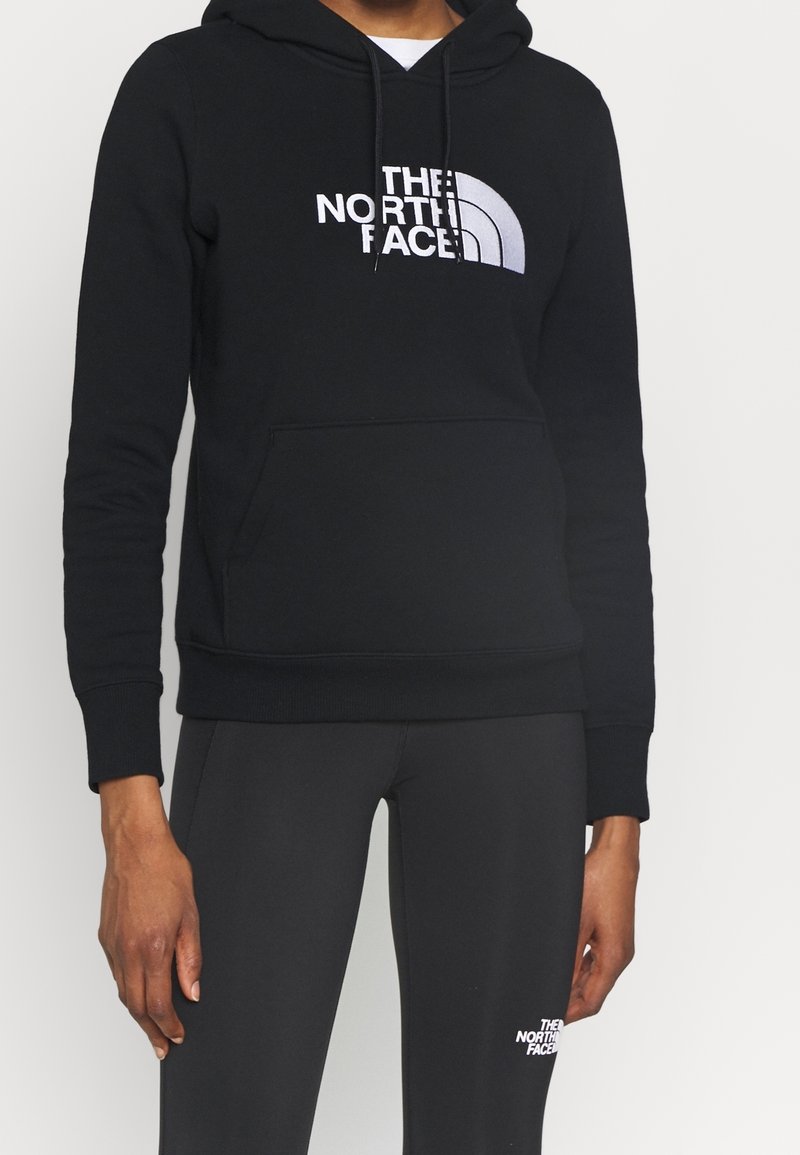 Personne portant un sweat à capuche noir The North Face avec logo sur la poitrine et un pantalon noir assorti avec logo près de la cuisse gauche.