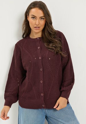 Kvinde iført en knaplukket, mørklilla kabelstrikket cardigan med lange ærmer og lyseblå jeans mod en ensfarvet baggrund.