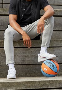 Witte sneakers met rode accenten, grijze sweatpants en een zwart shirt, zittend op houten treden naast een oranje en blauwe basketbal.