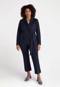 Laivastonsininen bleiseri, jossa on sidottu vyötärö, kaksi etutaskua, yhdistetty cropped-laivastonsinisiin housuihin ja sinisiin korkokenkiin. Tasainen kangas, jossa on rakenteellinen muotoilu.