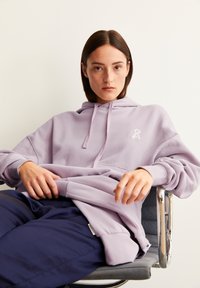 Helle lila Kapuzenjacke mit einer Vordertasche und Kordelzug, mit einem kleinen weißen Logo. Schwarze Hose, sitzend auf einem Stuhl mit minimalistischem Design.
