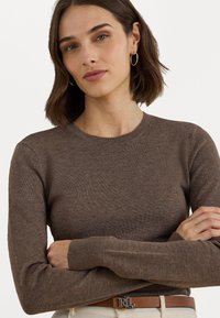 Bruine, gebreide trui met lange mouwen en een ronde hals; zachte textuur. Model draagt een lichte riem met een zilveren gesp, met gekruiste armen.