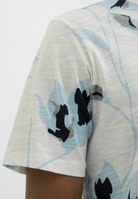 Camisa de manga corta de color claro con un patrón floral en tonos azules, negros y grises. Tejido suave y texturizado con un corte relajado.