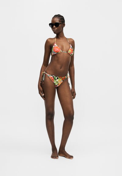 Γυναίκα στέκεται ξυπόλητη με πολύχρωμο φλοράλ μπικίνι και μαύρα γυαλιά ηλίου σε λευκό απλό φόντο.