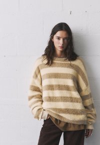 Raidallinen villapaita kermansävyisenä ja ruskeana, valmistettu pörröisestä materiaalista. Ominaisuuksia ovat leveä kaula-aukko ja rento istuvuus, yhdistettynä ruskeisiin samettihousuihin.