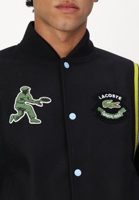 Veste de varsity noire avec des boutons-pression bleus, des bandes jaunes et des patchs brodés d'un joueur de tennis vert et du logo Lacoste.
