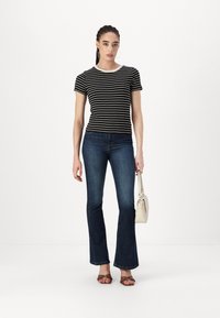 Chemise à rayures noires et blanches à manches courtes, jeans évasés bleu foncé, sandales brunes à lanières et un petit sac à main blanc.