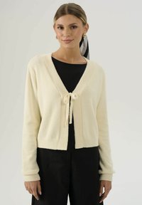 Cardigan corto color crema in un morbido tessuto testurizzato. Caratterizzato da un collo a V, bordi a coste e una chiusura con lacci davanti.