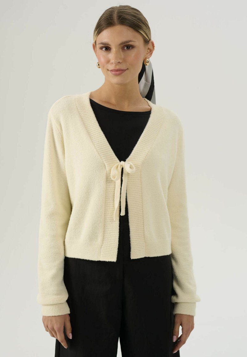 Cardigan corto color crema in un morbido tessuto testurizzato. Caratterizzato da un collo a V, bordi a coste e una chiusura con lacci davanti.