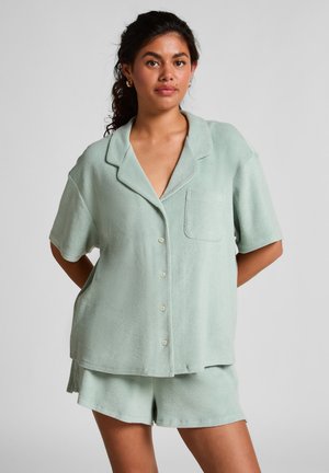 Vrouw met donker krullend haar, gekleed in een lichtgroene korte mouwen overhemd met knopen en bijpassende shorts, tegen een effen grijze achtergrond.