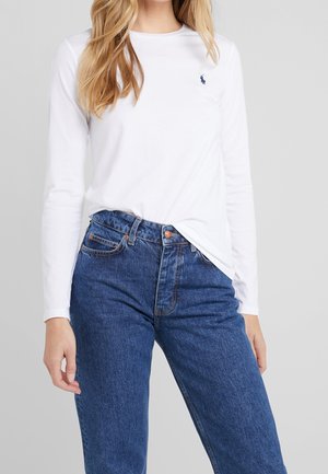 Femme portant une chemise blanche à manches longues avec un petit logo bleu marine sur la poitrine, assortie à un jean bleu en denim taille haute, sur un fond gris uni.