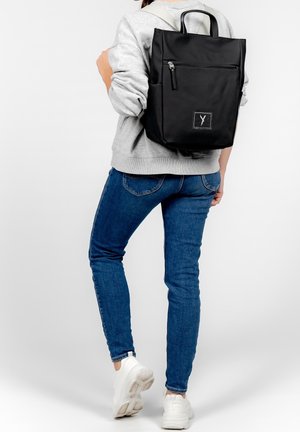 Personne portant un jean bleu, un sweat-shirt gris, des baskets blanches, transportant un sac à dos noir rectangulaire avec une poche avant zippée et des poignées supérieures.