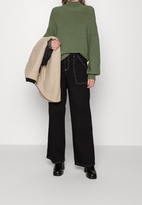 Zielony, prążkowany sweter z wysokim kołnierzem, zestawiony z czarnymi szerokimi spodniami z kontrastującym białym przeszyciem. Beżowa kurtka przewieszona przez ramię.