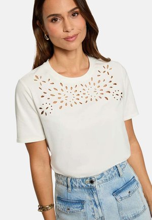 Femme portant un t-shirt blanc à manches courtes avec des motifs floraux ajourés sur la poitrine et un jean denim bleu clair taille haute.