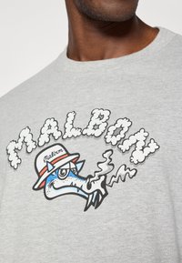 Γκρίζα μπλούζα με στάμπα ενός καρτούν δράκου π�ου καπνίζει, με το κείμενο "MALBON" σε γραμματοσειρά που θυμίζει σύννεφα, διακοσμημένη με κόκκινα και μπλε στοιχεία.