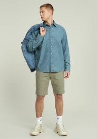 Camicia di jeans di un blu chiaro, allacciata, abbinata a pantaloni corti khaki. Il modello indossa calzini bianchi e sneaker beige, tenendo una giacca di jeans.