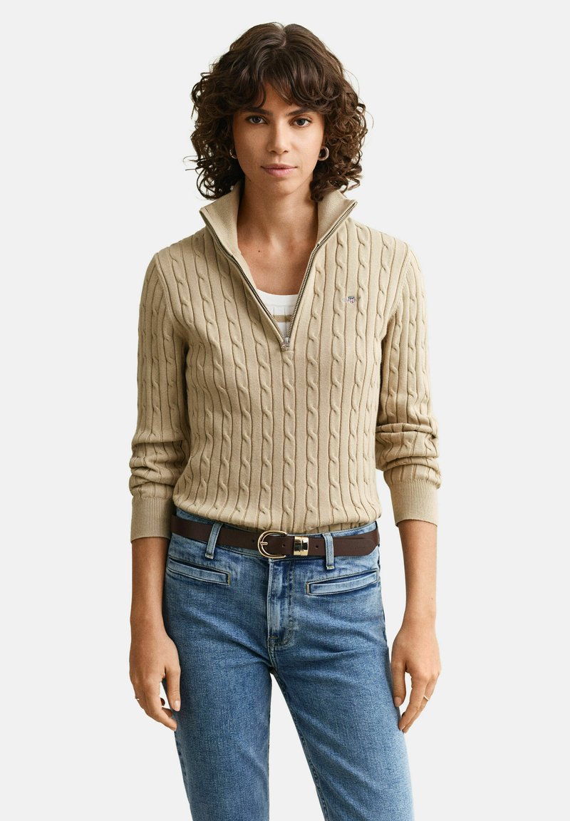 Femme aux cheveux bouclés portant un pull beige en maille torsadée à fermeture éclair à quart, sur un haut blanc, jean bleu et ceinture marron, debout devant un fond uni.