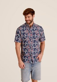Korte mouwen shirt in marineblauw met een bladerenpatroon in groen en rood. Heeft een button-down kraag en een relaxed fit. Gecombineerd met denim shorts.