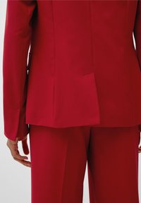 Costume rouge composé d'un blazer structuré avec une fente et d'un pantalon ajusté. Le matériau a une texture lisse. Mains positionnées près de la taille.