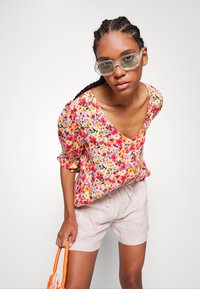 Blusa com padrão floral, com decote em V, mangas bufantes curtas e barra franzida. Combinada com calções de linho claros e uma bolsa de cor laranja.