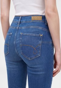 Gros plan d'une personne portant un jean denim bleu avec des poches arrière et une étiquette de marque marron clair sur la ceinture, associé à un haut blanc.