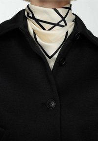 Czarny płaszcz z zapięciem na guzik ma kremowy szalik z czarnymi geometrycznymi liniami. Tkanina wydaje się gładka i uporządkowana.