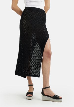 Donna che indossa una gonna midi in pizzo nero con spacco laterale, top bianco e sandali neri con plateau e cinturini alla caviglia, in piedi su sfondo bianco.